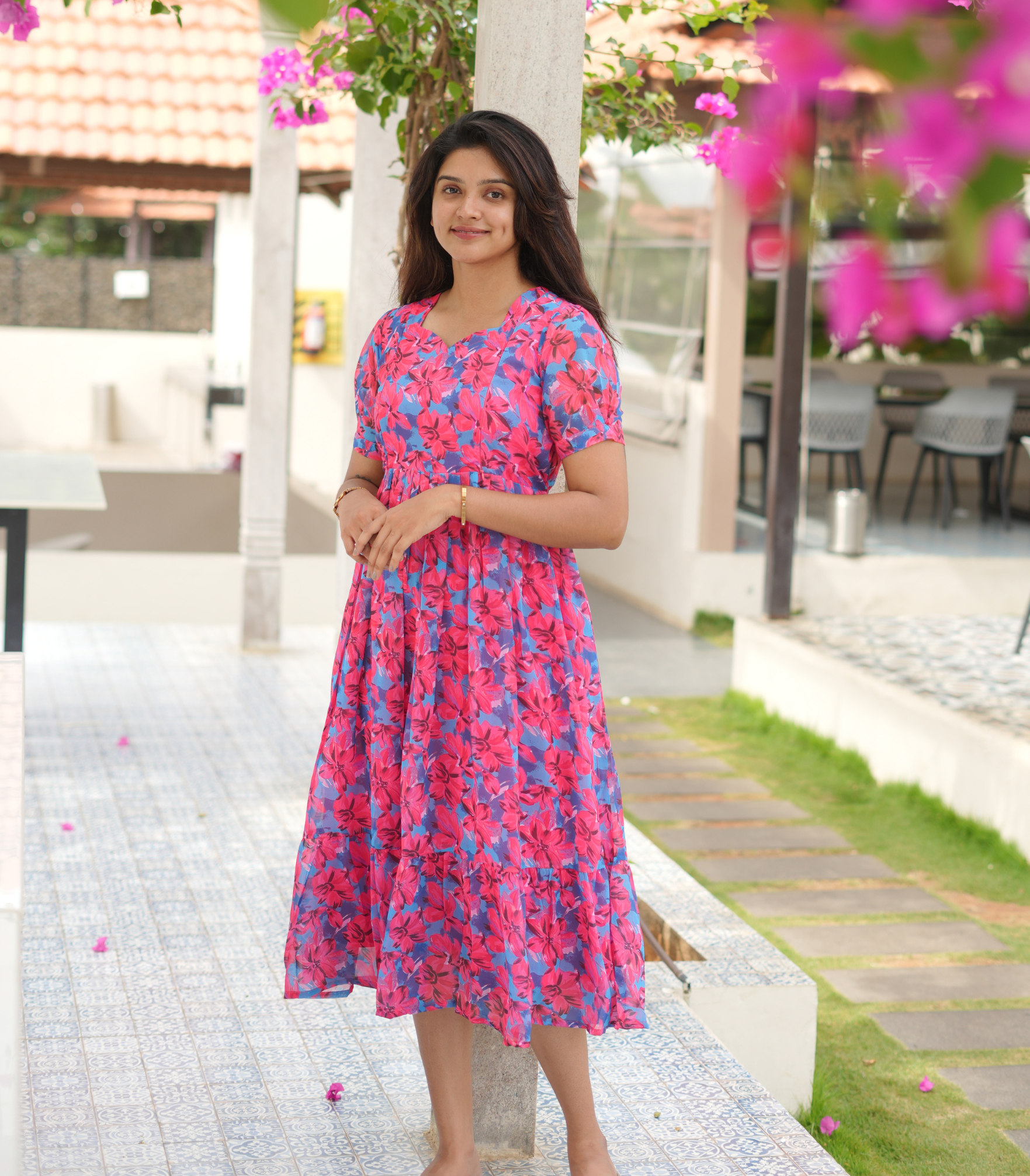 Woman in a pink floral dress standing in a garden with pink flowers and white columns.
