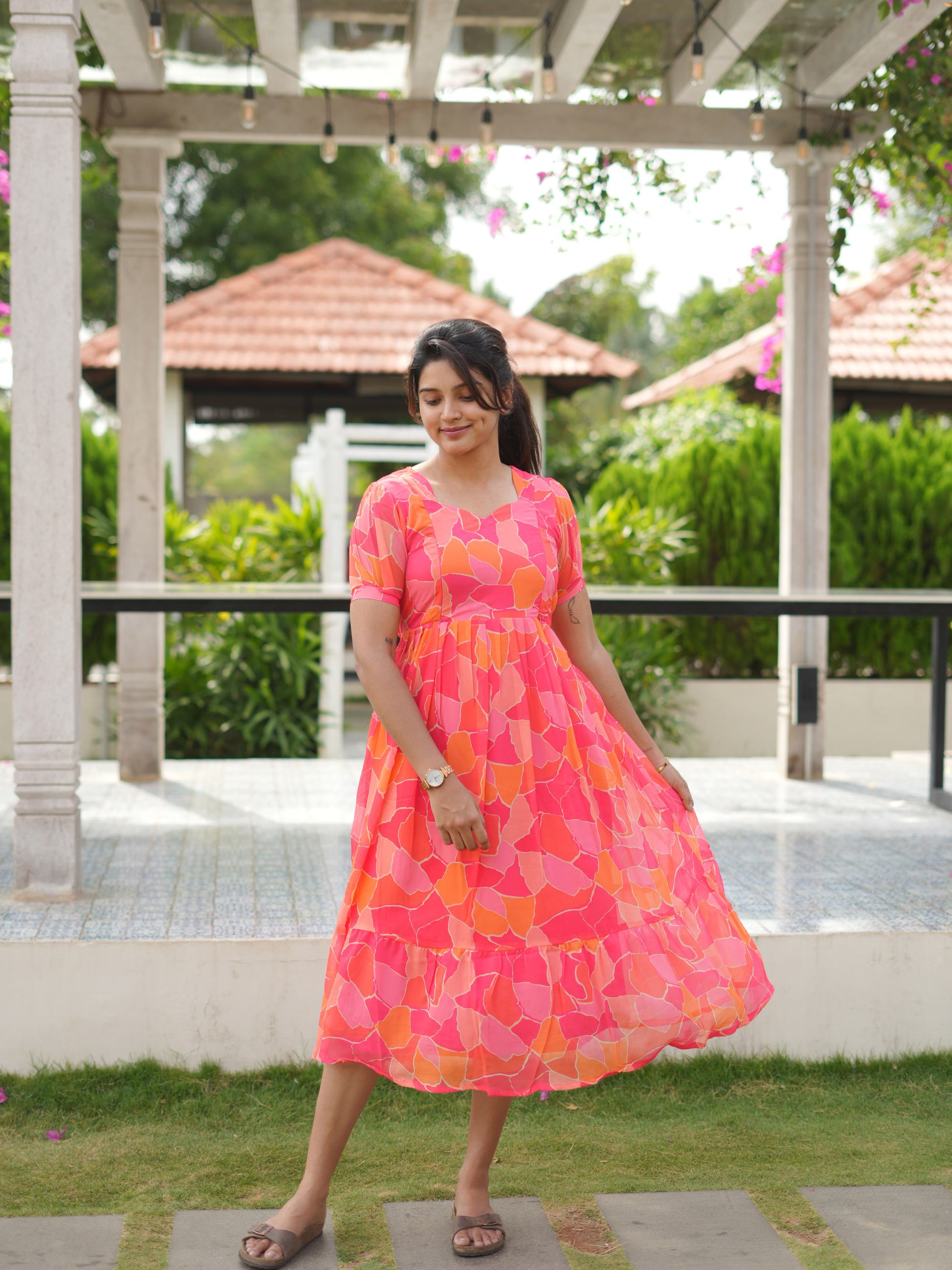 Woman in a colorful dress standing outdoors with greenery and a gazebo in the background