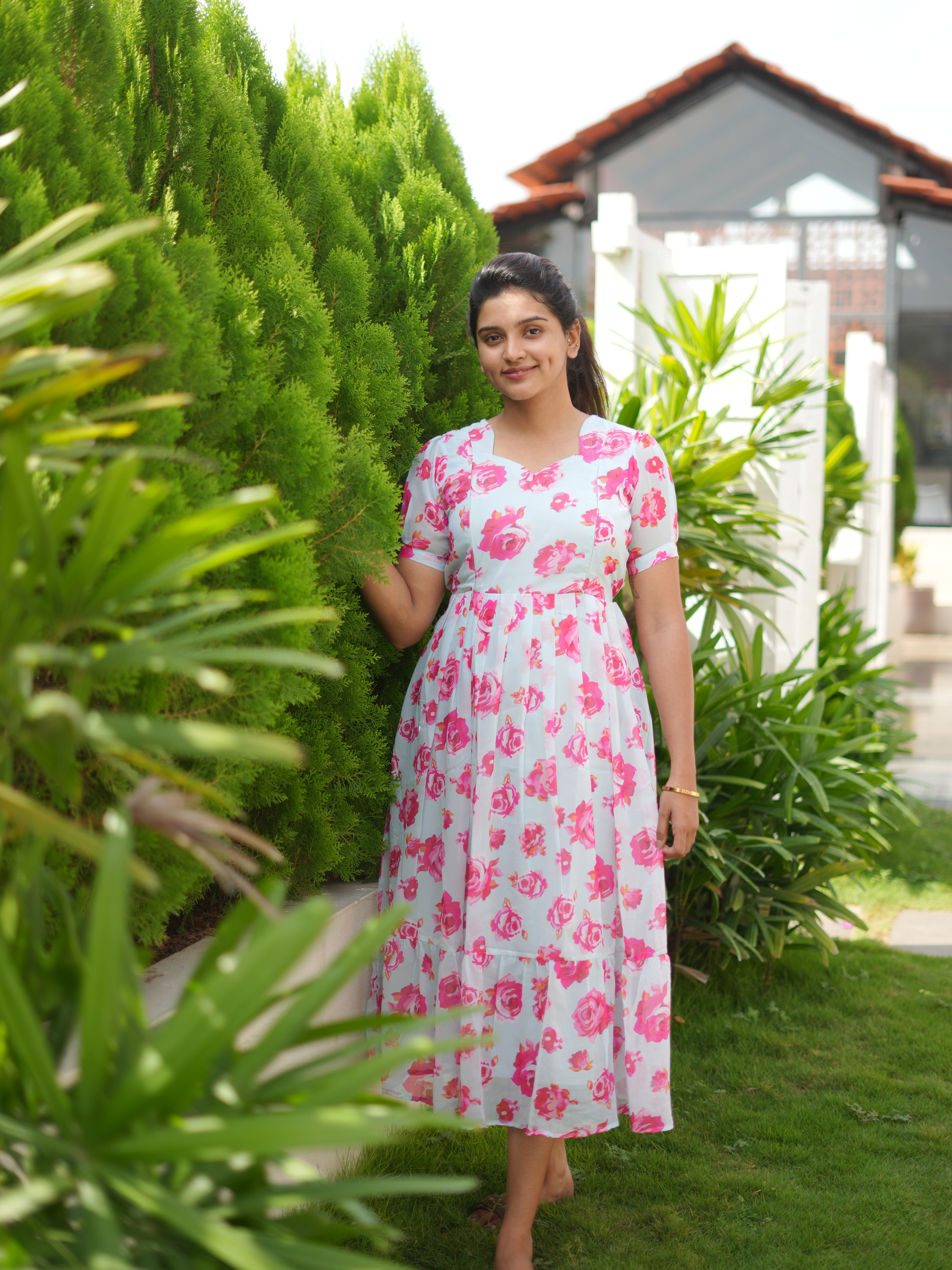 Woman in a floral dress standing in a garden with greenery and a house in the background