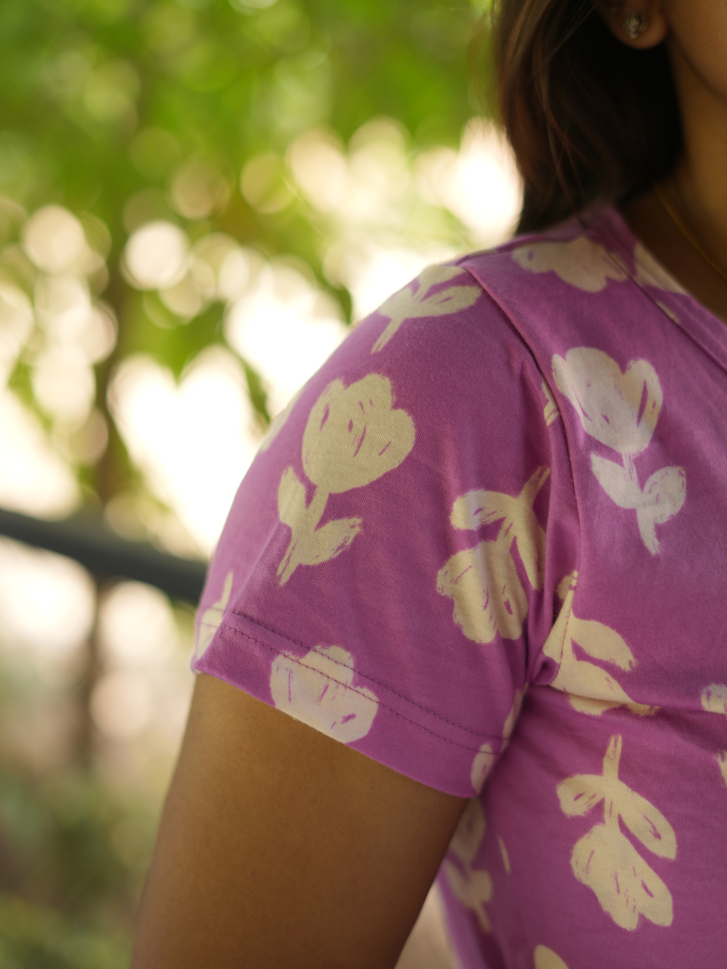 Woman wearing a purple shirt with white floral patterns, standing outdoors with greenery in the background.