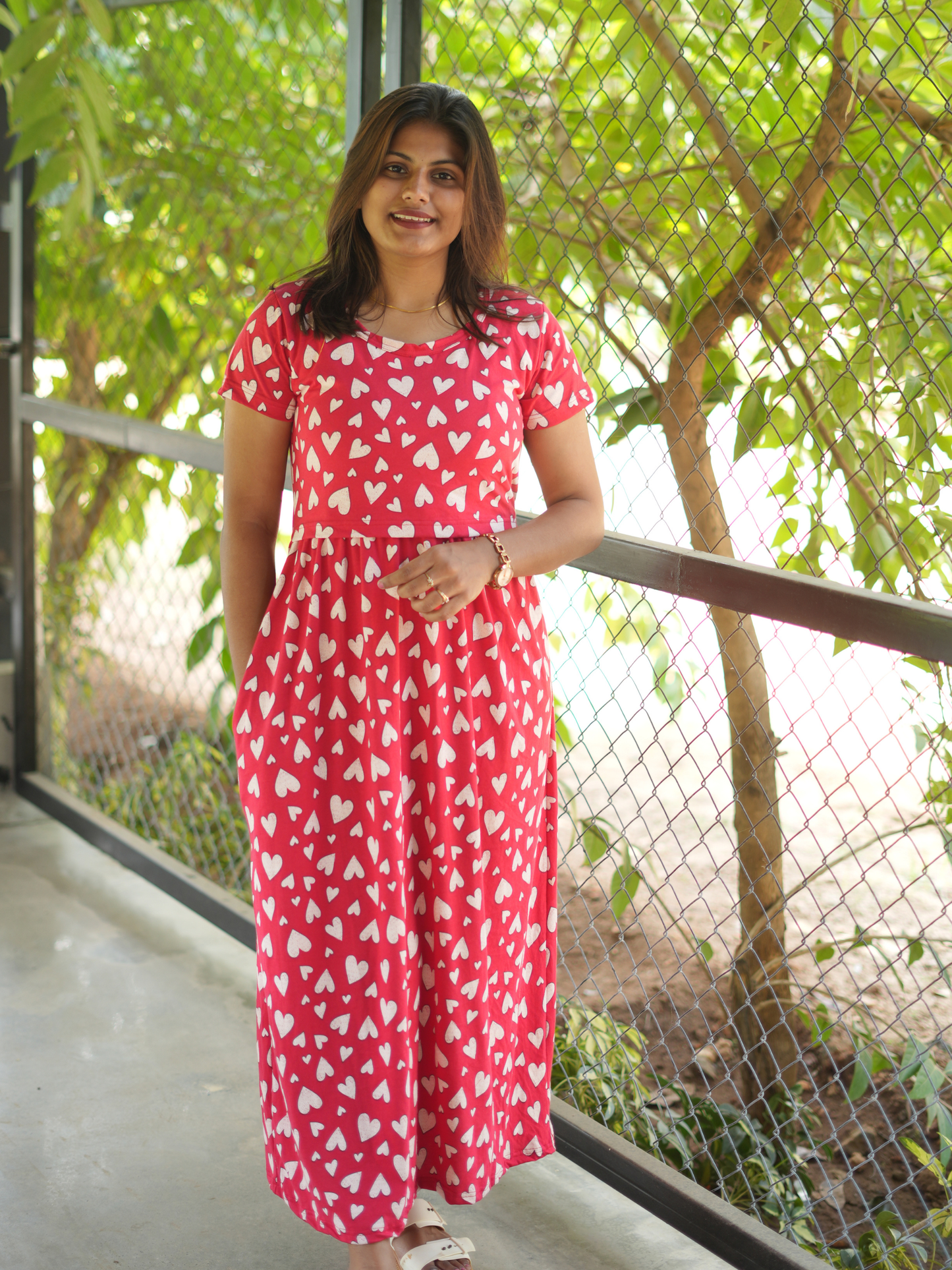 Woman wearing a red dress with white patterns standing outdoors with greenery in the background