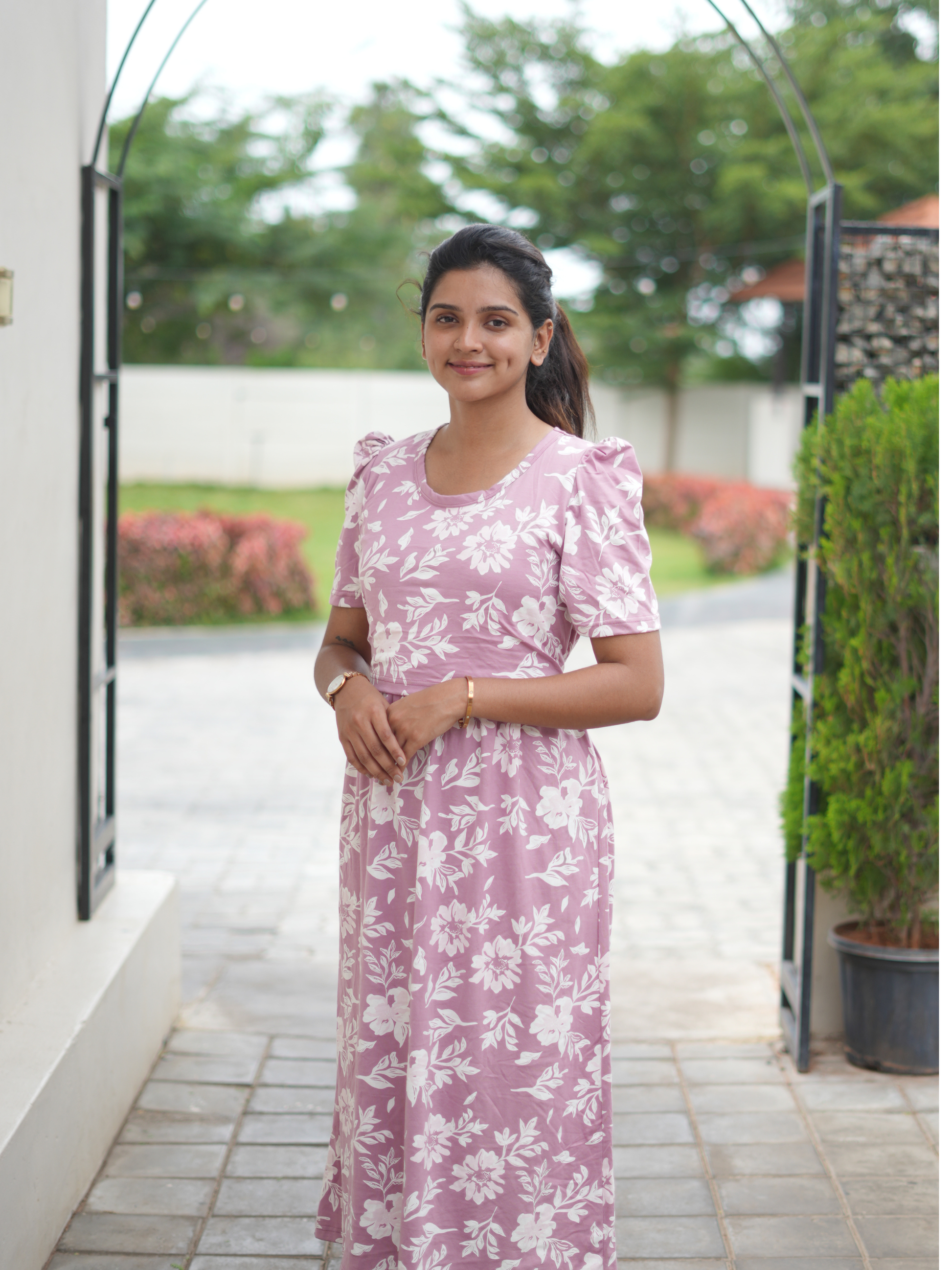 Woman in a pink floral dress standing outdoors with greenery in the background