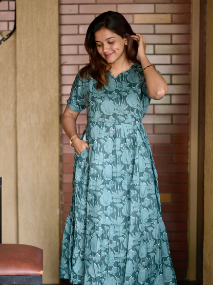 A woman wearing a green floral maxi dress with a V-neckline, featuring visible zippers and a pocket on the right side.