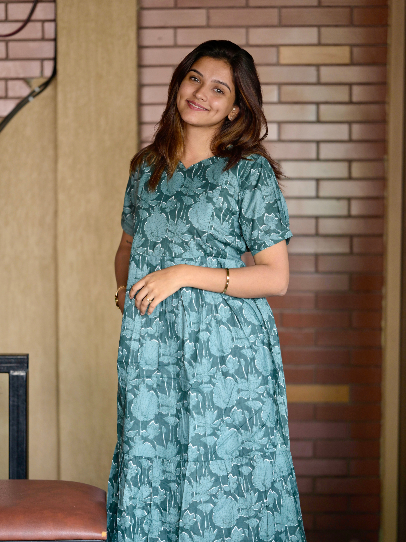 A woman wearing a green floral maxi dress with a V-neckline, featuring visible zippers and a pocket on the right side.