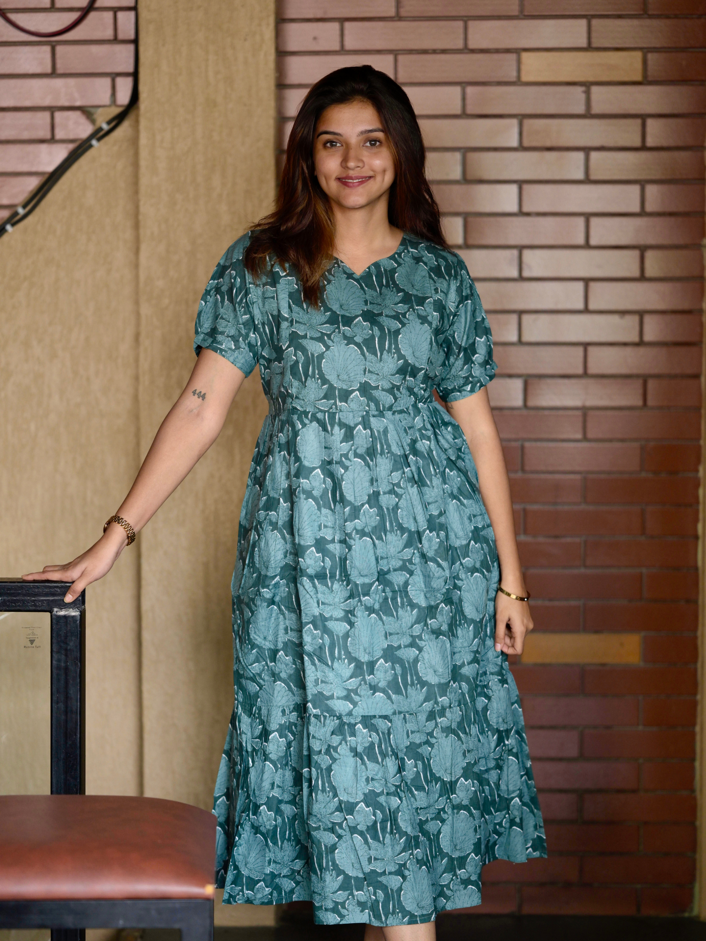 A woman wearing a green floral maxi dress with a V-neckline, featuring visible zippers and a pocket on the right side.