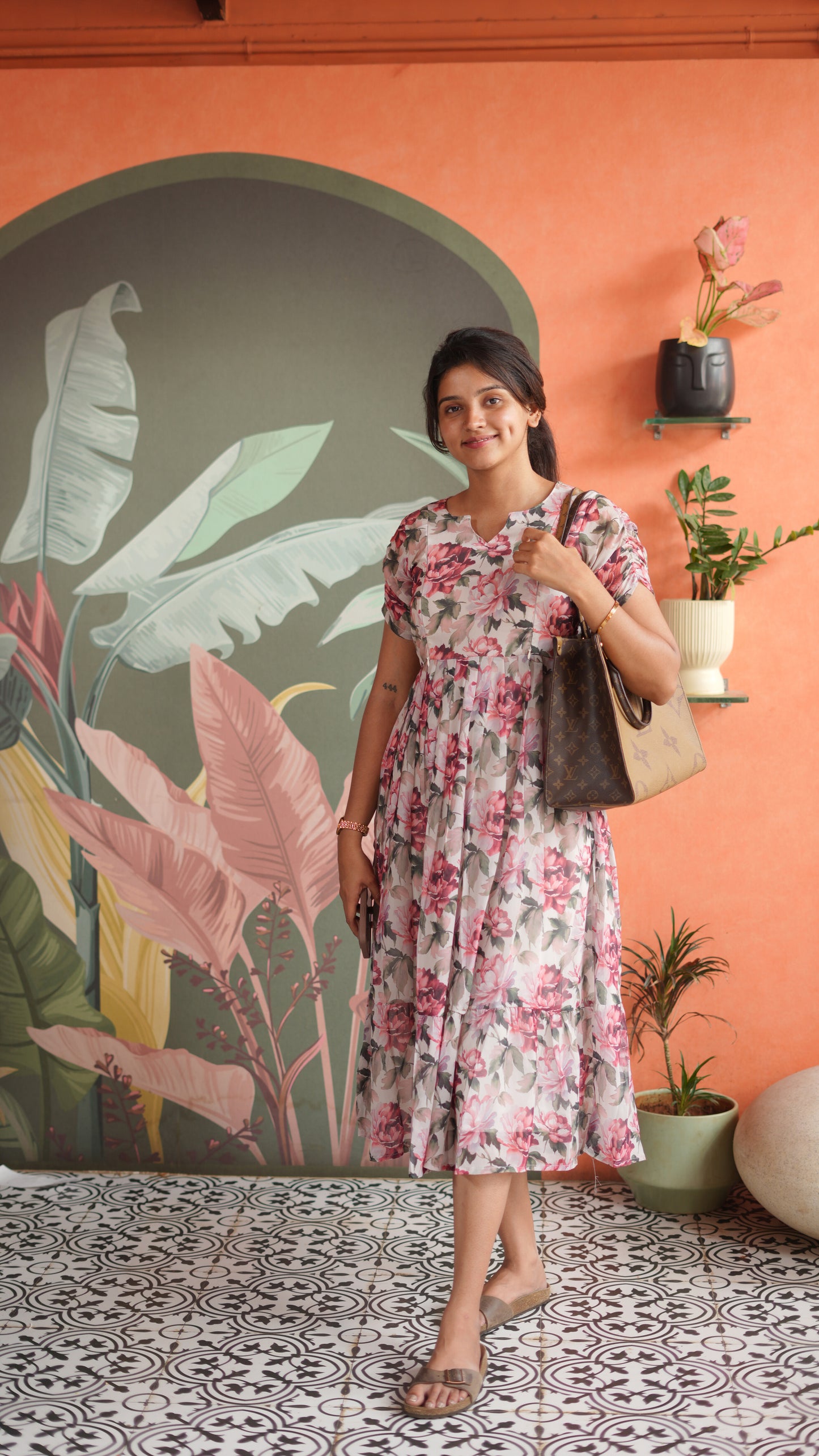 Woman in a floral dress holding a handbag against an orange wall with plant decorations