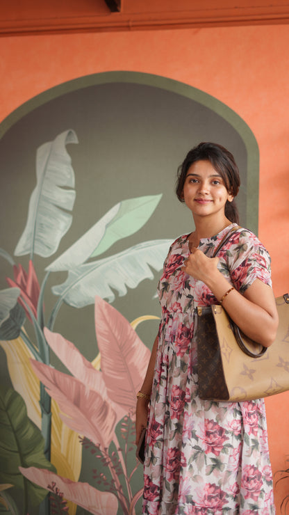 Woman in a floral dress holding a beige handbag against a wall with a colorful leaf design.