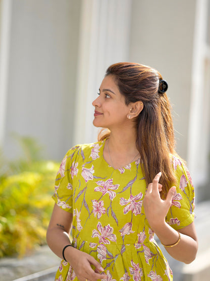 A woman standing on a pathway wearing a neon green maxi dress with a floral pattern, featuring adjustable straps and two visible zippers.