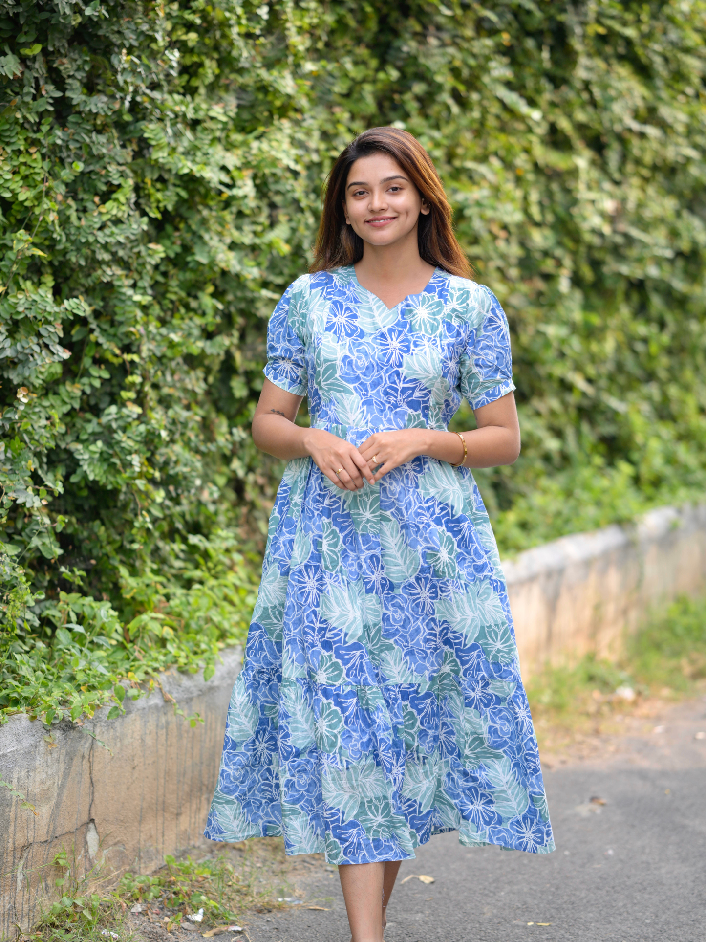A woman standing by a green bush wearing a blue and white floral maxi dress with three-quarter sleeves and a zipper at the back.
