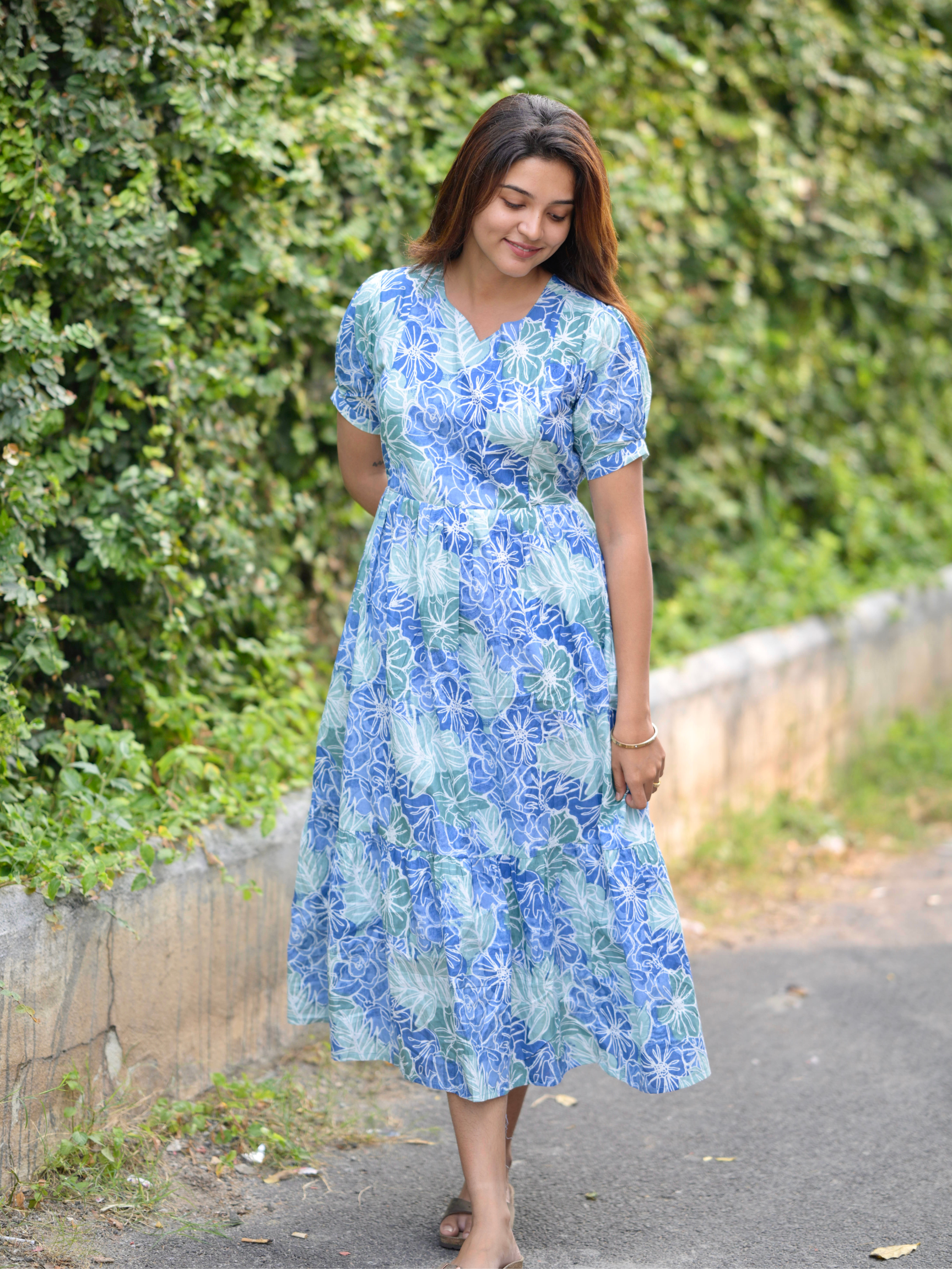 A woman standing by a green bush wearing a blue and white floral maxi dress with three-quarter sleeves and a zipper at the back.